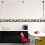 Modern kitchen with black base cabinets, white countertop, sink on the left, and a coffee maker on the counter; decorative tile border runs along the wall, plus a white table with a bowl of apples and colorful chairs nearby.