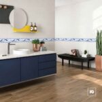 Cenefa cerámica f delfín 8 × 25 cm decorativa-tiles-2-ok | Plomería García Modern bathroom with navy vanity, white countertop sink, round mirror, and a yellow shelf holding two bottles; bench against tiled wall with a purse and small boxes; tall plant nearby.