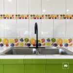 Double-basin stainless steel sink with a tall black gooseneck faucet, white tiled wall with a colorful fruit-pattern backsplash, and lime-green base cabinets.