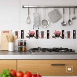 Modern kitchen with a gas stove, hanging utensils on a rail, and a spice jar rack above a gray countertop; fresh tomatoes in the foreground.