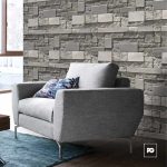 Light gray upholstered armchair with a floral pillow in a room with a textured stone accent wall and a blue rug nearby, bright window to the side.