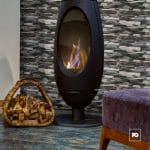 Contemporary freestanding fireplace with a glass front burning logs, next to a basket of firewood and a purple upholstered stool in a living room.