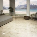 Modern bedroom with marble tile floor and a beach view through floor-to-ceiling windows; bed on the left and a white lounge chair on the right.