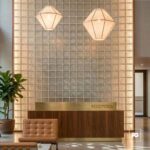 Hotel reception desk in front of a glass-block wall with two geometric pendant lights overhead, and a potted plant nearby.