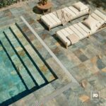 Mosaico alberca sage-30x61-bali-cenezzia-2-ok | Plomería García Overhead view of a sunlit pool deck with blue-green tiled pool, steps, and two cream cushioned lounge chairs with pillows and a beige throw. A round wooden table with a plant sits nearby.