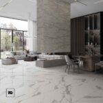 Piso porcelánico tipo mármol blanco brillante colossale 80 × 160 cm-tendenzza-2-ok | Plomería García Open, airy living-dining space with marble floors, a large sectional sofa, a central linear fireplace, and a wooden dining table, illuminated by large windows showing greenery; fg logo bottom-left.
